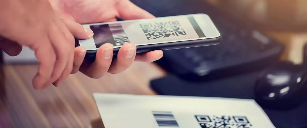 Person holding a smartphone displaying a 2D QR code and traditional 1D barcode above product packaging featuring both barcode formats.