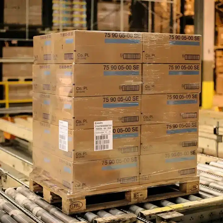 Stacked cardboard boxes secured on a wooden pallet with barcode labels applied, moving along a conveyor system in a warehouse.