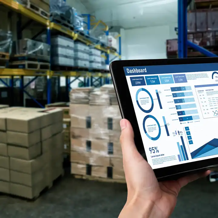 Person holding a tablet displaying production analytics and performance charts inside a warehouse with palletized inventory and storage racks.