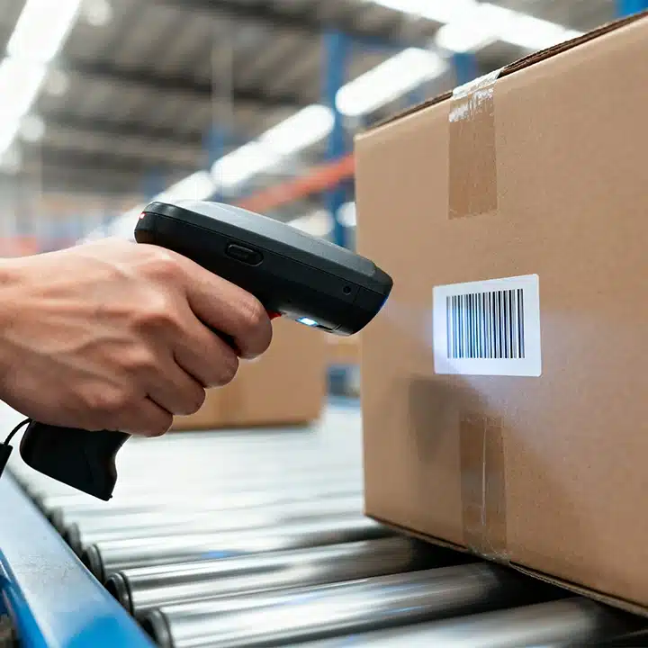 Warehouse worker using a handheld barcode scanner to scan a shipping label on a cardboard box moving along a conveyor.