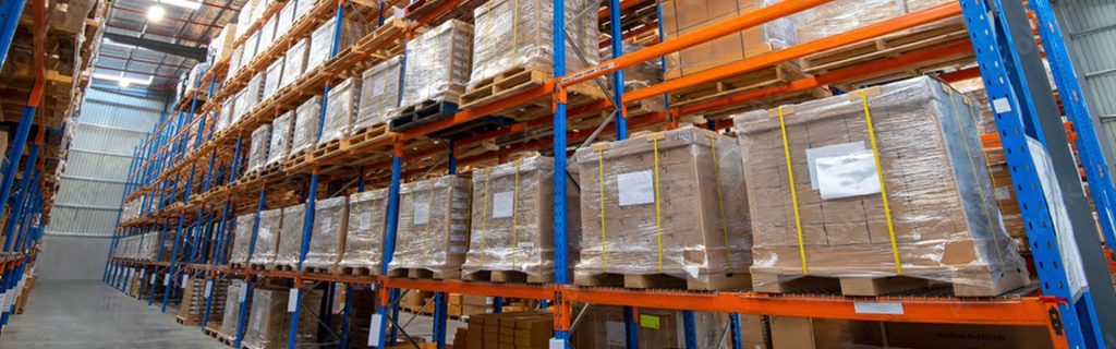 Rows of shrink-wrapped pallets containing cardboard boxes stored on industrial warehouse shelving for shipping and distribution.